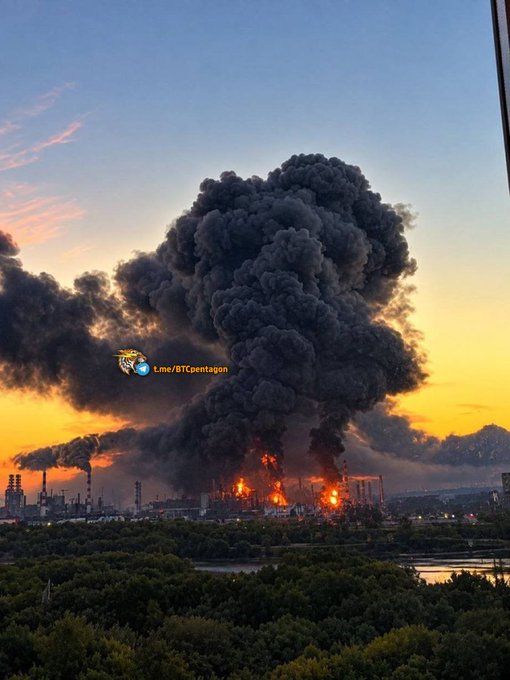 A dramatic photograph of a massive explosion at an industrial facility with a huge black smoke plume rising against an orange sunset sky, watermarked "Line/BTCpentagon".