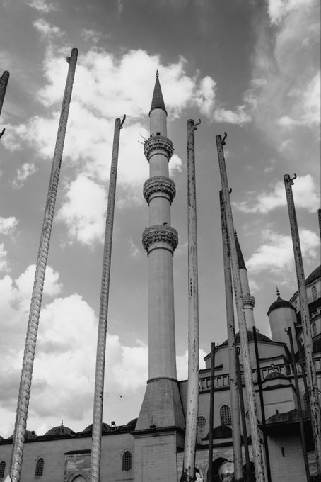 A black-and-white low-angle photograph of a mosque minaret and dome surrounded by tall poles against a cloudy sky.