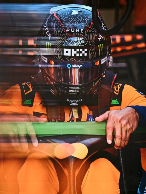 A McLaren F1 driver in an orange racing suit sits in the cockpit wearing a black helmet branded with "OKX," "Android," and "Pure Electric" logos, with the post referencing Monza likely hinting at a moment before the Italian Grand Prix. The reflective, slightly blurred shot through the garage glass captures the quiet preparation moment behind the scenes.