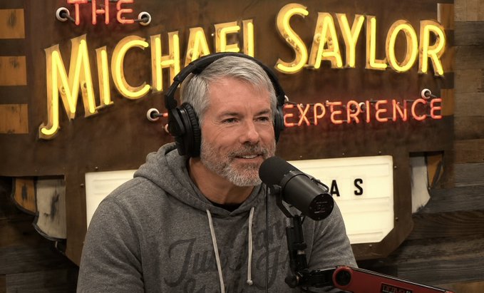 A man with gray hair and a beard wearing headphones and a gray hoodie, sitting in front of a microphone at 'The Michael Saylor Experience' podcast studio with a neon sign in the background.