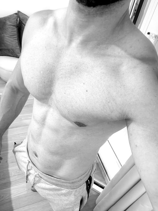 A black and white mirror selfie showing a man's bare torso, highlighting his chest and abdominal muscles while wearing light-colored shorts.