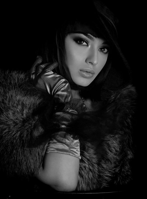 A dramatic black-and-white portrait of a woman wearing a dark hood and fur stole, with satin gloves resting near her face as she gazes intensely at the camera.