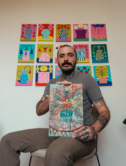A bearded, tattooed man sits holding a colorful artwork depicting a vase of flowers, with a grid of 15 similar vibrant vase-themed paintings displayed on the wall behind him.