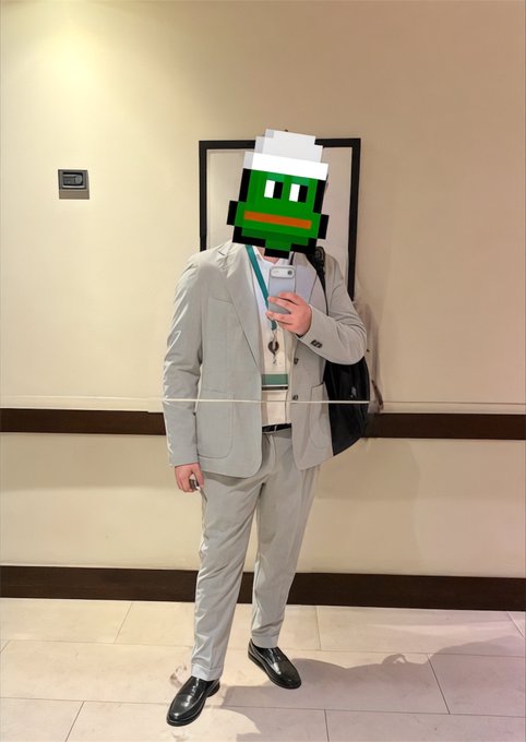 A mirror selfie of a man in a light gray suit with a lanyard, with a Pepe the Frog meme sticker covering his face, standing in a hallway.