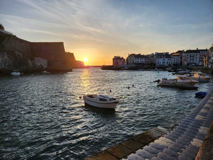 A scenic photograph captures a sunset over a coastal harbor town, with boats floating on the water and buildings lining the shore.