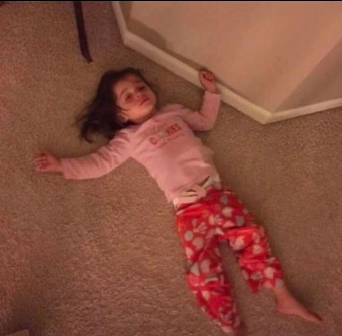 A humorous photo of a young girl lying on a carpeted floor against a wall, smiling while wearing pink pajamas and red floral pants.