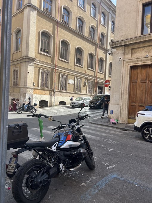 A blue and white BMW motorcycle is parked on a cobblestone street in a European city, with classic buildings, cars, and pedestrians visible in the background.