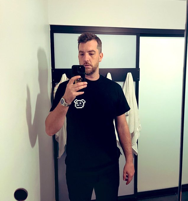 A man takes a mirror selfie with a smartphone, wearing a black t-shirt featuring a white pixel-art character design and a silver wristwatch.