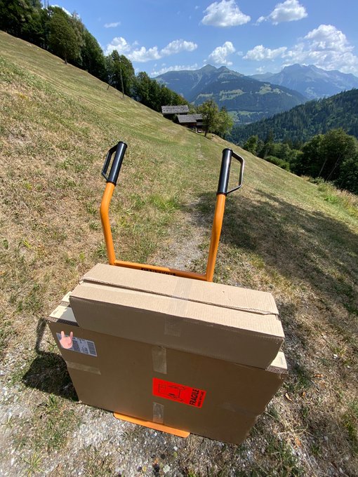 A cardboard box being transported via an orange hand truck on a grassy mountain trail, with alpine peaks and a wooden cabin visible in the background.