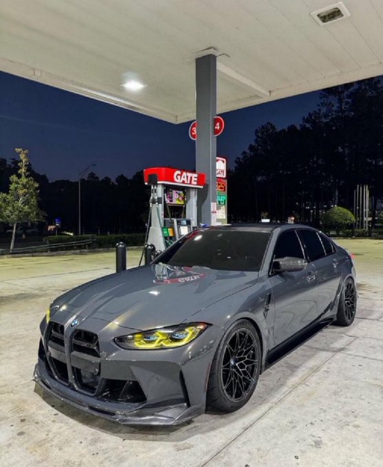 A gray BMW sedan parked at a gas station at night, featuring an aggressive front splitter, black wheels, and yellow headlights.
