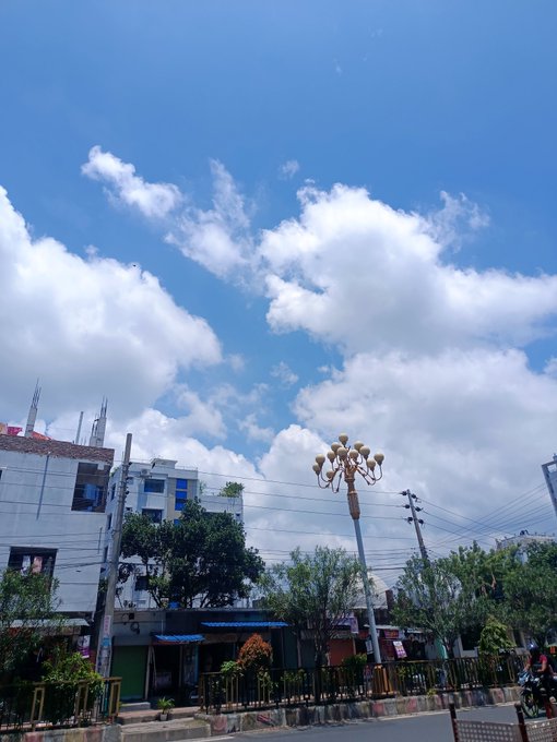 A daytime outdoor scene showing a bright blue sky with scattered white clouds over a street with buildings, trees, and a decorative lamppost.