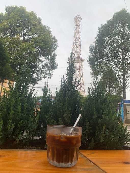 An iced coffee in a glass sits on a wooden table with a view of a red and white communication tower behind some trees.