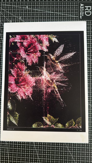 A piece of artwork or print featuring pink flowers and a star-like burst on a dark background rests on a green cutting mat with grid lines.