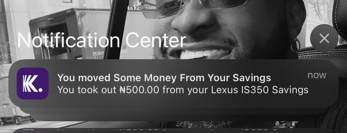 A black and white image of a smiling man overlaid with a 'Notification Center' alert from 'K.' stating, 'You moved Some Money From Your Savings. You took out ₦500.00 from your Lexus IS350 Savings.'