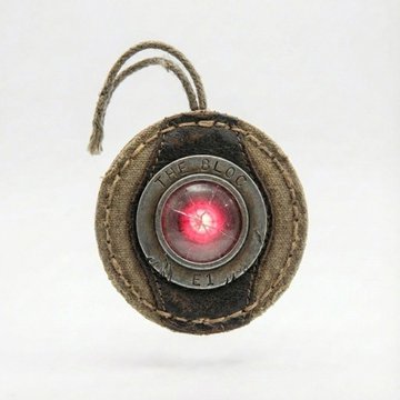 A small round button or badge with the text 'THE BLOG' around its edge, featuring a glowing red center light, displayed on a leather backing with a string loop at the top.