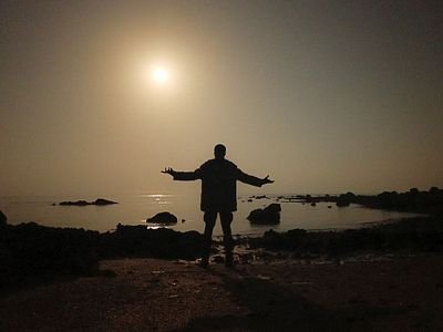 A silhouetted figure stands with arms outstretched facing a bright sun over water and rocky shoreline at dusk.