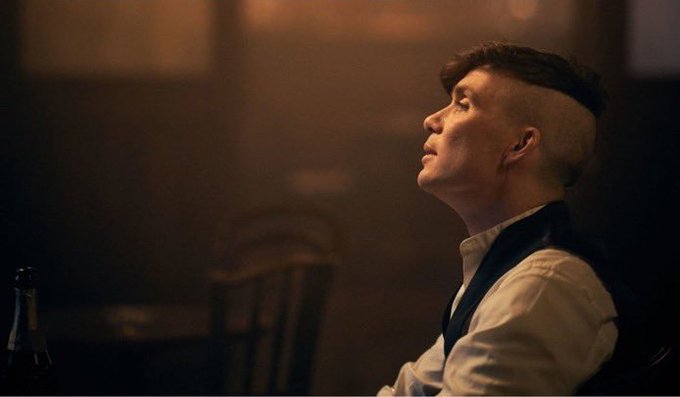A man with a shaved-side hairstyle, wearing a white shirt and dark vest, sits in a dimly lit room looking upwards with his eyes closed.