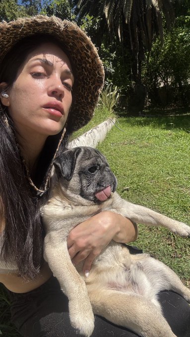 A woman wearing a leopard-print bucket hat poses outdoors in a sunny garden, holding a panting pug dog in her arms.