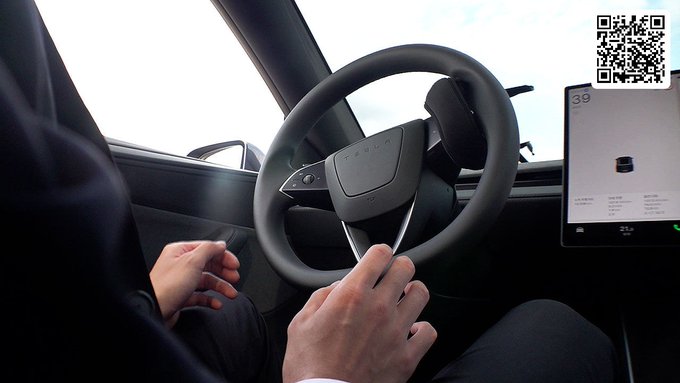 An interior view of a Tesla vehicle showing a person's hands near the distinctive rectangular steering wheel, with the central touchscreen displaying a vehicle interface and a QR code in the upper right corner.