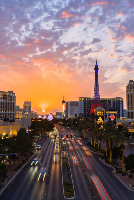 A sunset view of the Las Vegas Strip featuring the Eiffel Tower replica of the Paris Las Vegas hotel, with light trails from traffic on the road below.