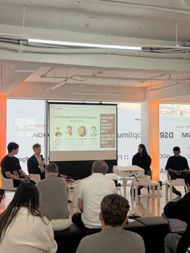 A panel discussion at a corporate event with multiple speakers seated on stage in front of a presentation screen, viewed from the audience perspective.