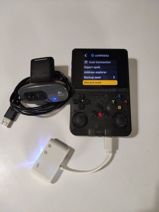 A SeedSigner Bitcoin hardware wallet device displaying its menu options (Scan transaction, Export xpub, Address explorer, Backup seed, Discard seed) alongside a connected Logitech webcam with an illuminated blue LED and a white USB adapter cable, demonstrating the project's camera functionality.