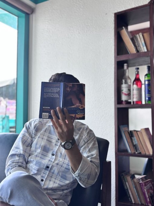 A person sitting in a chair by a window reading a book that obscures their face, wearing a plaid shirt and a wristwatch, with a bookshelf and bottles visible in the background.
