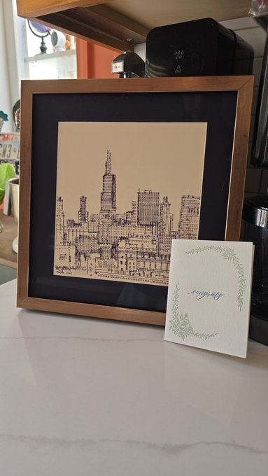 A framed artwork of a city skyline on a kitchen counter next to a sympathy card with green floral borders.