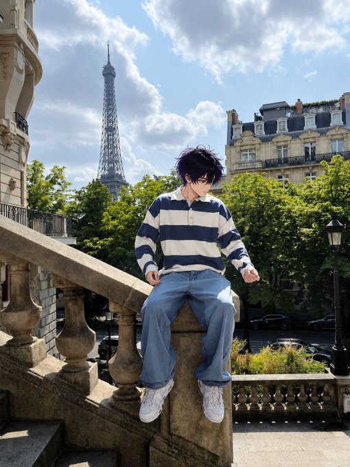 An anime-style illustration of a young man wearing a striped shirt and jeans, sitting on a stone balcony railing with the Eiffel Tower and Parisian buildings visible in the background.