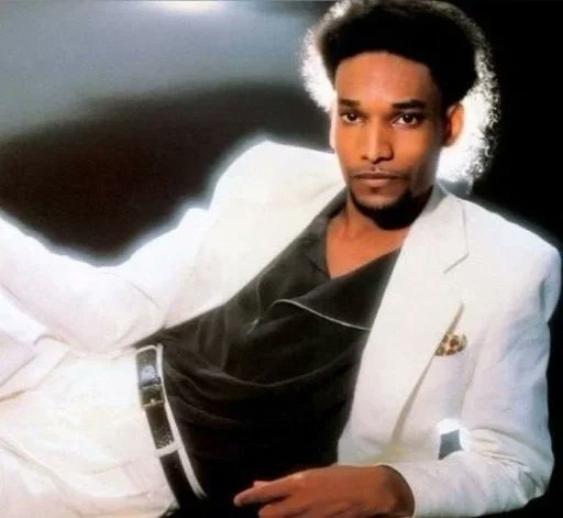 A vintage-style portrait photograph of a young man with an afro hairstyle and goatee, wearing a white blazer over a dark shirt.