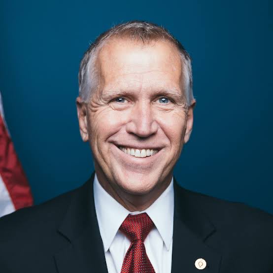 Senator Thom Tillis smiles in a formal portrait wearing a dark suit and red tie, with an American flag visible behind him. The image accompanies news about his procedural maneuvers regarding the Clarity Act, where he strategically switched his cloture vote to position himself on the prevailing side for future reconsideration.