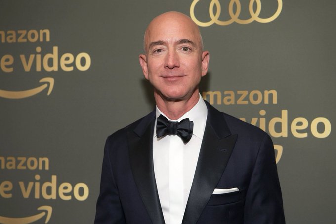 The image features a man wearing a suit and a tuxedo with a black bow tie. He appears to be smiling for the camera, posing against a backdrop of a green wall adorned with gold text. The caption above him reads "AMAZON IS NOW HIRING BITCOIN AND CRYPTO PROFESSIONALS UP TO $500,000 A YEAR THE PIVOT IS REAL". This suggests that the man might be involved in Bitcoin mining or cryptocurrency trading and could potentially earn a substantial income from these activities. The presence of this text above him adds context to his attire and reinforces the idea that he is part of an industry that has seen a rise in demand for skilled professionals in areas like digital currencies, blockchain technology, and financial innovation.