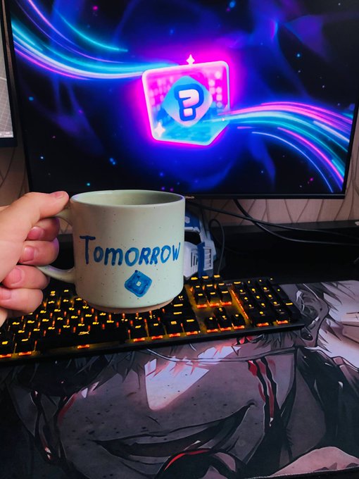 A hand holds a cream-colored mug labeled "Tomorrow" with a small blue question mark block illustration, positioned in front of a backlit mechanical keyboard with orange LEDs. In the background, a monitor displays a glowing blue question mark block icon against a vibrant purple and blue cosmic wallpaper.
