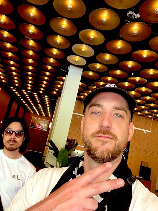A selfie-style photo of a bearded man wearing a black Adidas cap and white shirt, making a peace sign, with another man in sunglasses visible in the background under a ceiling covered in numerous golden disc lights.