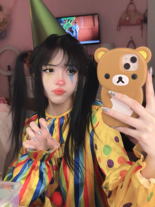 A young woman takes a mirror selfie wearing clown makeup with colorful teardrops, a party hat, and a yellow and striped clown costume, holding a Rilakkuma phone case.