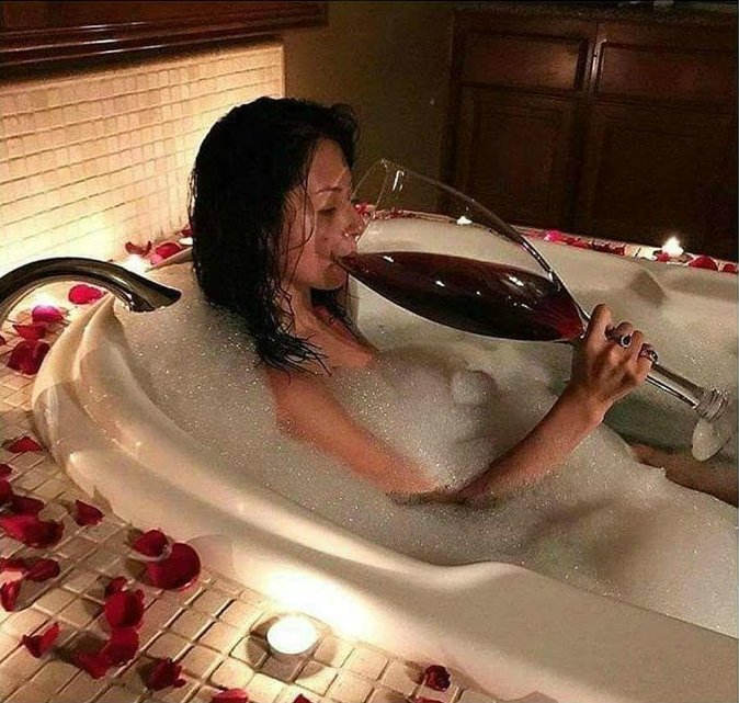 A woman relaxes in a bubble bath surrounded by rose petals, drinking red wine from an oversized novelty glass in a candlelit bathroom setting.