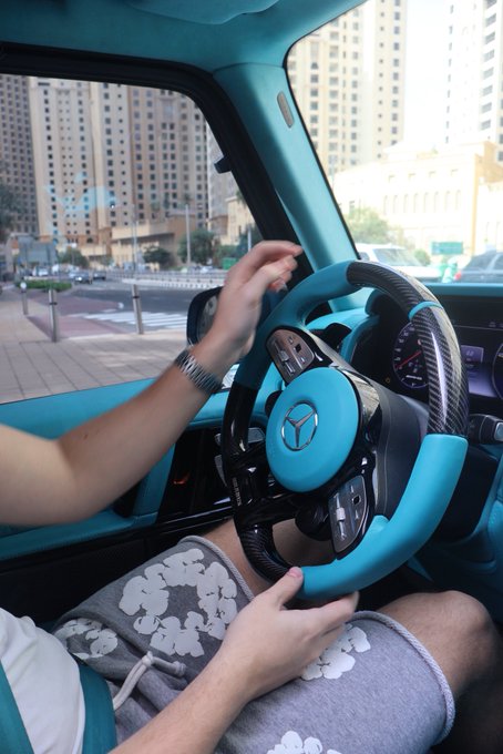 A person sits behind the wheel of a teal and black Mercedes-Benz, gripping the steering wheel in a brightly colored customized interior, with the caption "GM (get money)" playfully riffing on the iconic three-pointed star logo.