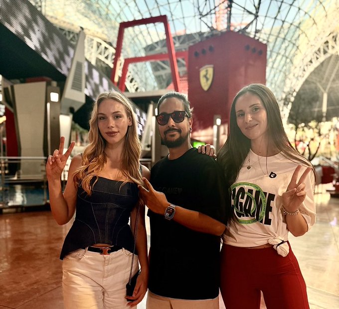 Three people pose together in what appears to be a Ferrari-themed venue, with a red structure and Ferrari shield logo visible in the background; the two women make peace signs while the man in the center wears sunglasses.
