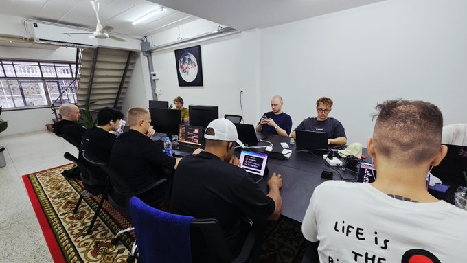 A group of people sit around a conference table working on laptops in an office space with a staircase and an astronaut artwork on the wall.