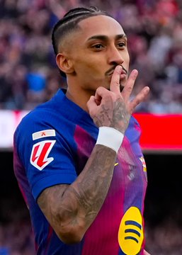 A photograph of FC Barcelona footballer Raphinha in the team's blue and red striped jersey, making a gesture with his fingers to his lips while on the pitch.