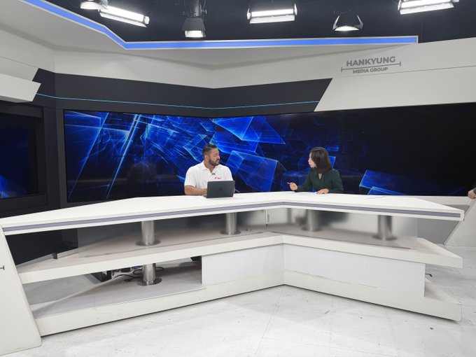 Two presenters, a man and a woman, seated at a modern white anchor desk in a broadcast studio for 'HANKYUNG MEDIA GROUP,' with a large blue geometric graphic on the LED screens behind them.