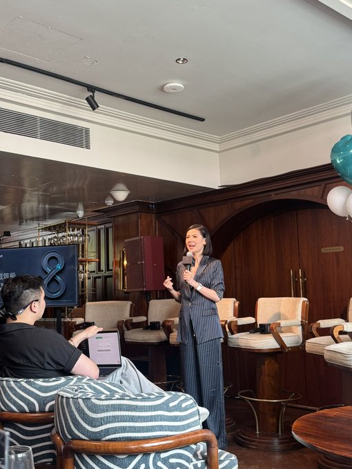 A woman in a striped suit speaks into a microphone at an indoor event, while a man in the foreground uses a laptop.
