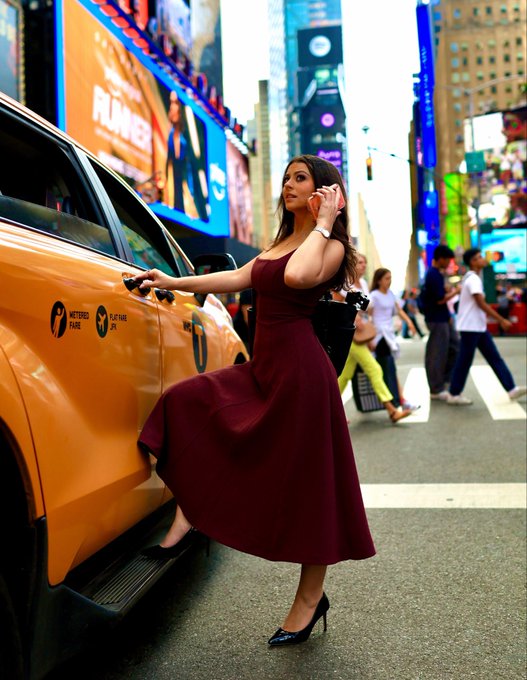 A woman in a maroon dress steps out of a yellow taxi in what appears to be Times Square, talking on her phone. The image thematically captures the post's message about Bitcoin reclaiming the $100k level—portraying confidence, style, and a return to the spotlight amid the bustling financial backdrop of NYC.