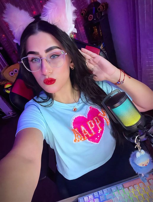 A young woman with dark hair and glasses wearing fluffy white ears and a light blue 'HAPPY' t-shirt, posing next to a colorful RGB microphone and keyboard.