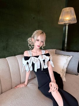 A young woman with styled blonde curls and red lipstick poses on a beige sofa, wearing a black off-shoulder dress with a light blue bow detail, set against a dark green wall beside a tall lamp. The Japanese hashtag "#自撮り界隈" translates to "#selfieworld" or "#selfiecommunity," fitting the image's posed, portrait-style self-portrait aesthetic.