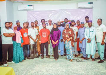 A group photo of around twenty people posing together indoors, with some wearing traditional Nigerian attire, in front of a banner, with two air conditioning units visible on the wall above.