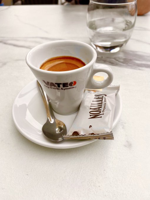 A small cup of espresso with a 'NATCIO' branded saucer and a 'NOMALLE' sugar packet sitting on a marble table next to a water glass.