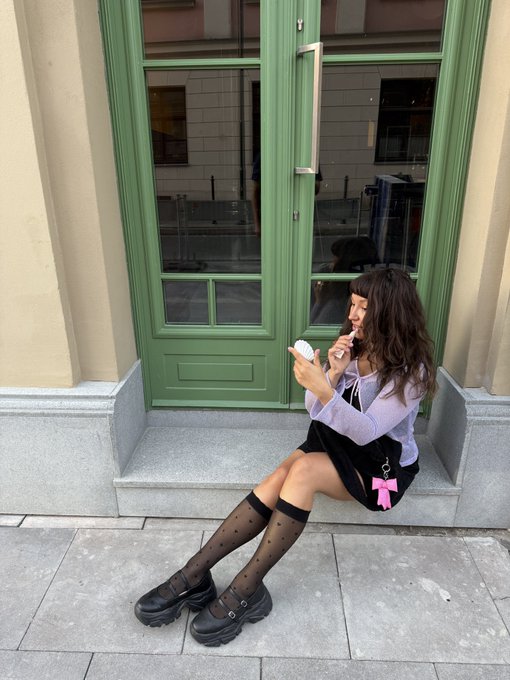 A young woman with long dark hair sits on a step in front of a green door, wearing a purple top, black skirt, polka dot knee-high socks, and chunky black shoes while looking at a compact mirror.