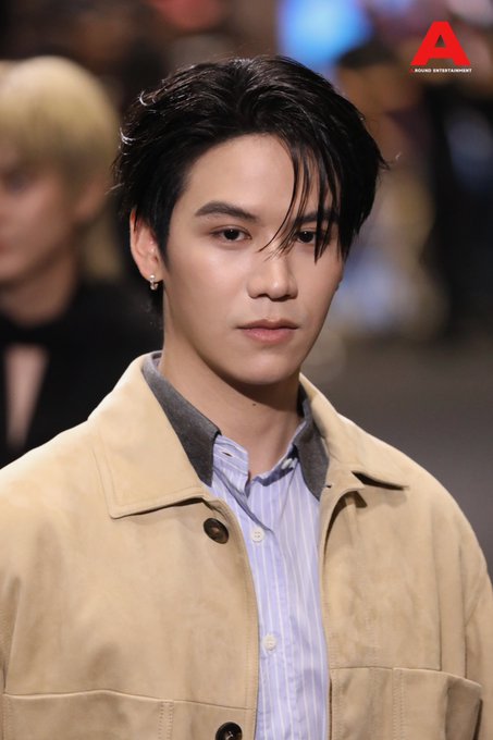 A young man with dark, swept-back hair and an earring poses on a red carpet, wearing a beige jacket over a striped shirt. The image relates to the post about "สตางค์ กิตติภพ" (Satang Kittiphop) attending the 'Mchoice & Mint Awards 2026' event, with the red "A" logo and text "ASIA ENTERTAINMENT" visible in the top right corner of the photo.