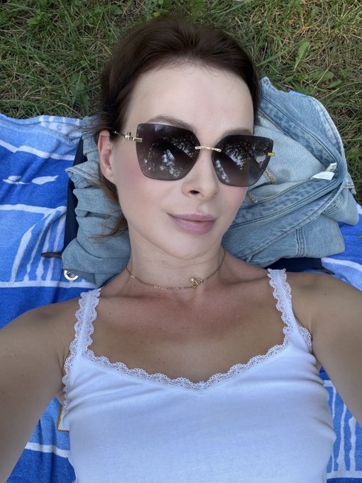 A woman with long brown hair wearing large sunglasses, a white lace-trimmed tank top, and a delicate gold necklace lies on a blue and white striped blanket on grass.